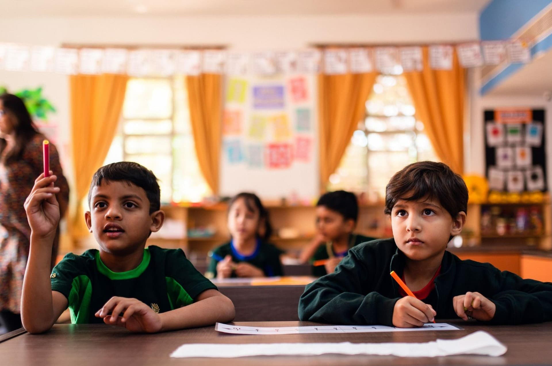 Dos niños con uniforme verde atentos en el salón de clases; uno de ellos levanta la mano con un lápiz para participar.