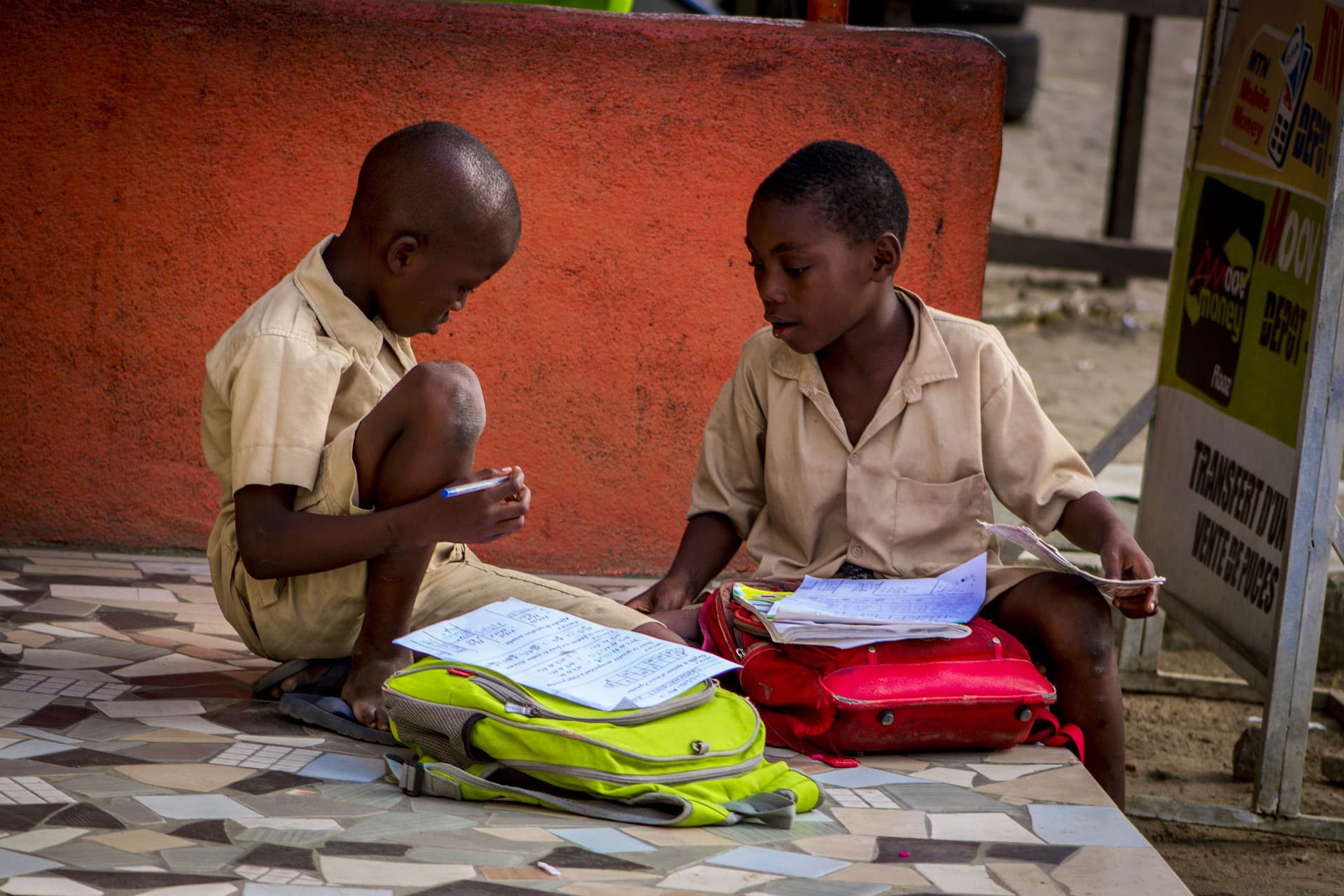 Dos niños con uniforme estudian juntos sentados en el suelo, con mochilas, cuadernos y hojas de ejercicios.