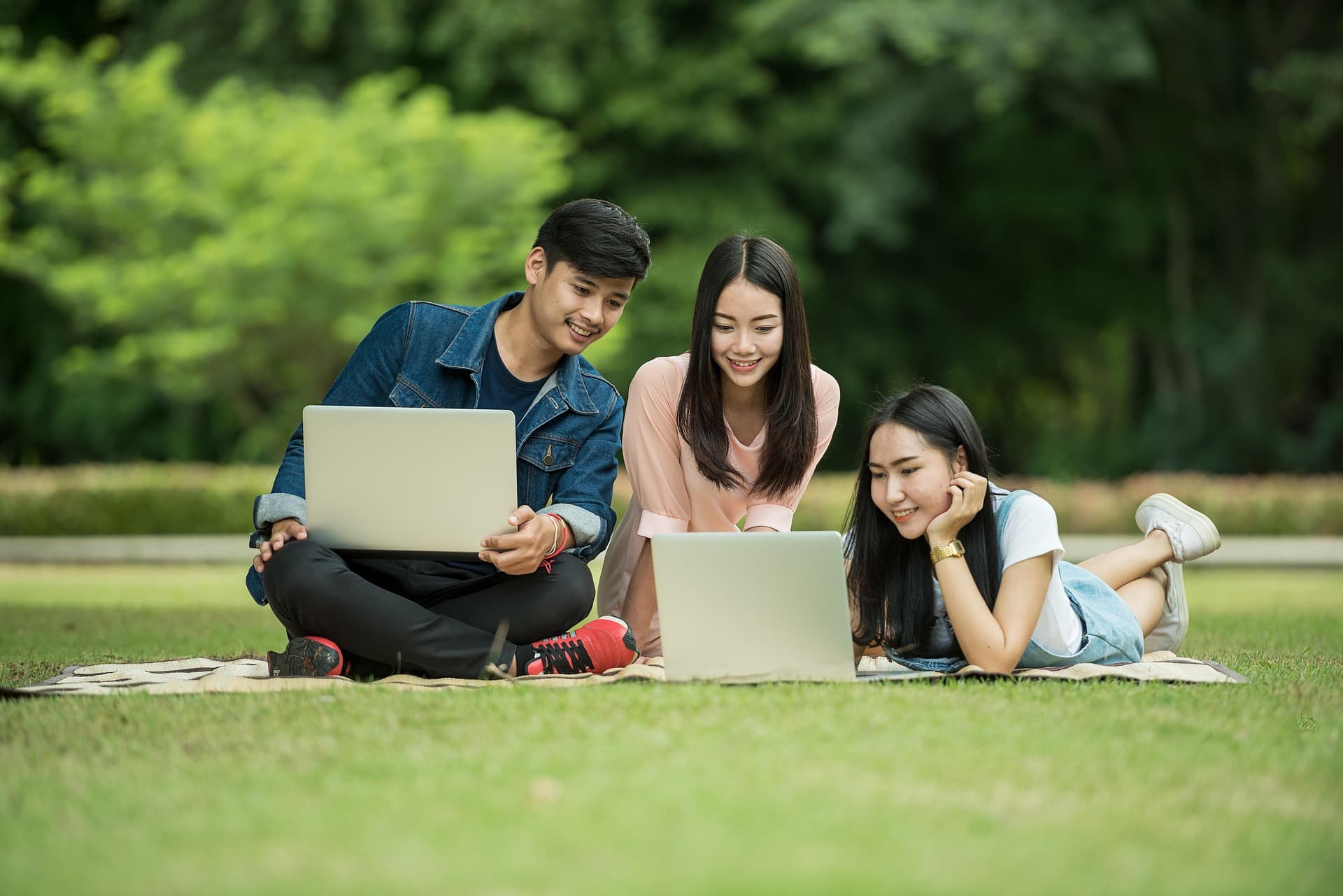 Tres jóvenes sonrientes trabajan juntos con computadoras portátiles sobre el césped de un parque.